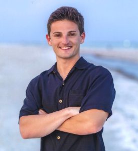 Young man with short brown hair smiling at the camera, wearing a dark navy button-down shirt on a beach.